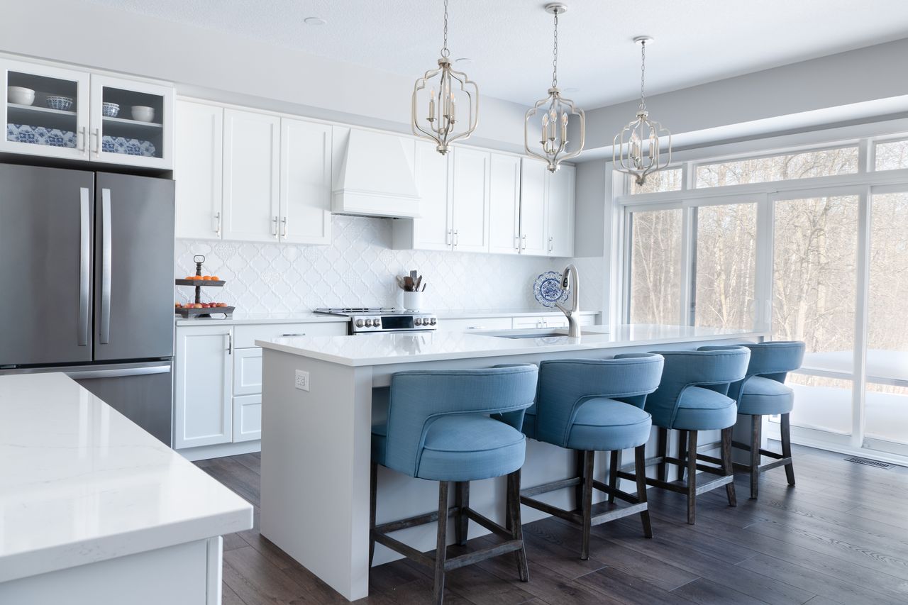 Elegant modern kitchen featuring white cabinets, blue bar stools, and large windows overlooking a snowy outdoors.